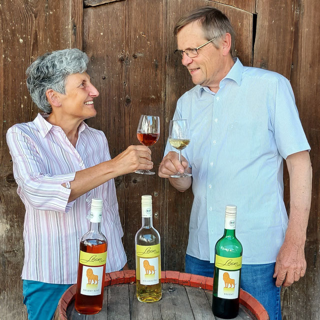 Frau und Mann stehen an einem Fass, auf dem Weinflaschen stehen, und prosten sich mit einem Weinglas zu