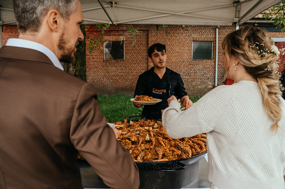 bruidegom en bruid schuiven aan bij een grote pan paëlla-huwelijksfotografie-trouwreportage-byFotografils-IlseWagemakers-Fotograaf gevestigd in Essen