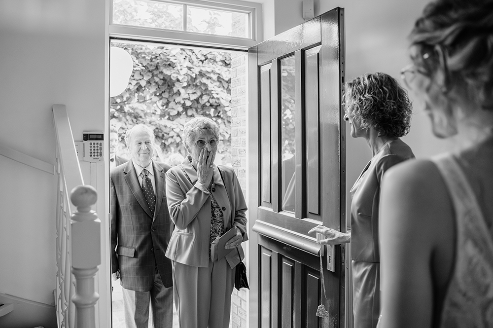 grootmoeder in de deuropening kijkt verbaasd en geëmotioneerd als de bruid verschijnt-huwelijksfotografie-trouwreportage-byFotografils-IlseWagemakers-Fotograaf gevestigd in Essen