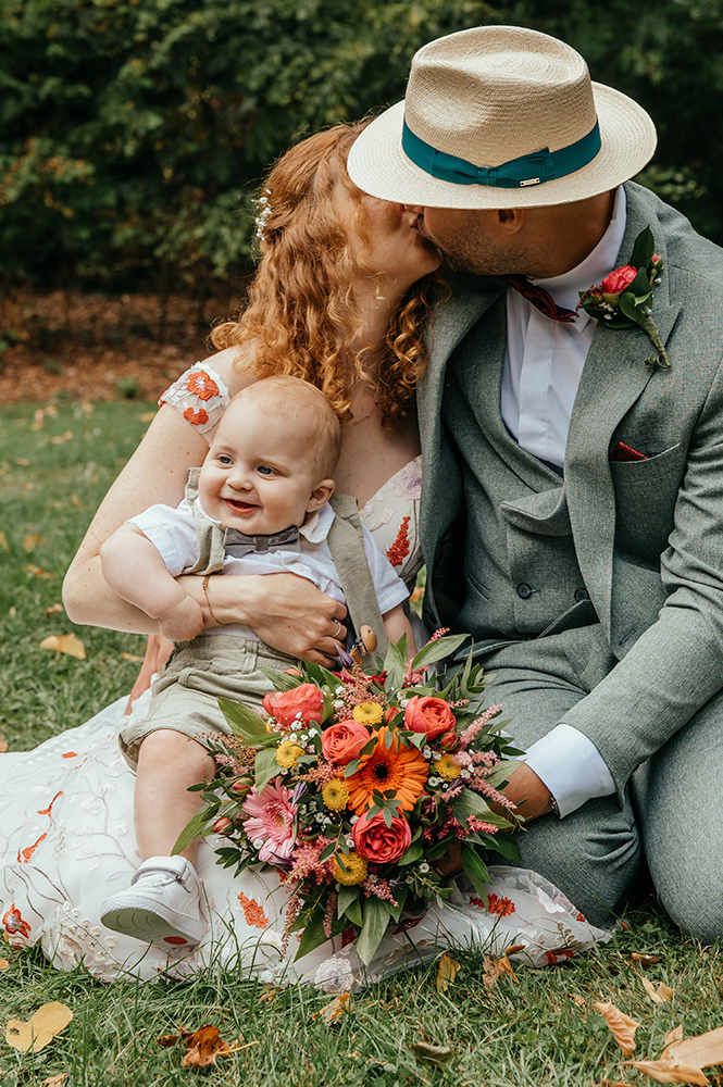 bruid en bruidegom kussen verstopt achter het hoedje van de bruidegom, het bruidskindje zit terwijl op hun schoot te lachen-huwelijksfotografie-trouwreportage-byFotografils-IlseWagemakers-Fotograaf gevestigd in Essen