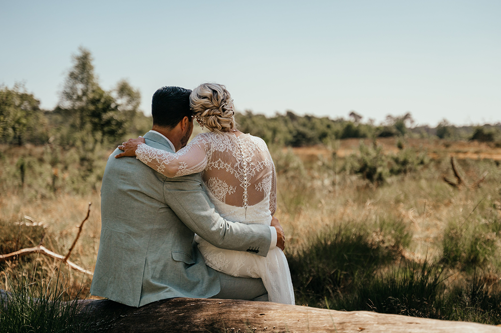 foto genomen vanop de rug; bruid en bruidegom zitten al knuffelend te genieten van het uitzicht van de Kalmthoutse heide-huwelijksfotografie-trouwreportage-byFotografils-IlseWagemakers-Fotograaf gevestigd in Essen