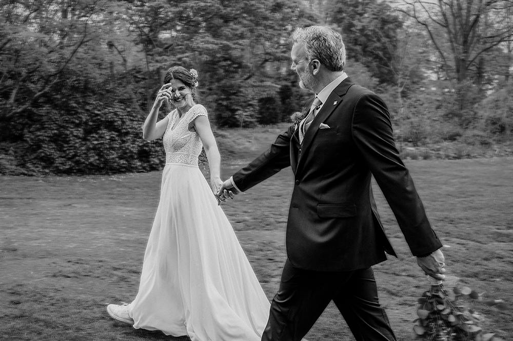 bruid trekt de bruidegom al lachend mee op wandel en strijkt daarbij met haar vrije hand door haar haren-huwelijksfotografie-trouwreportage-byFotografils-IlseWagemakers-Fotograaf gevestigd in Essen
