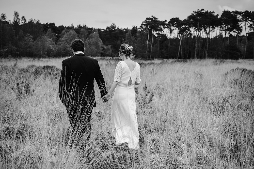 zwart wit foto van bruispaar op de rug gefotografeerd, wandelend door het lange gras in de Kalmthoutse heide-huwelijksfotografie-trouwreportage-byFotografils-IlseWagemakers