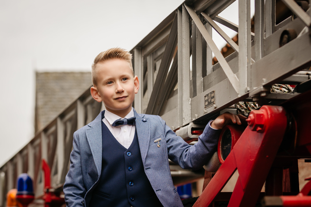 jongen in net kostuum leunt op een grote brandweerladder en tuurt glimlachend in de verte-communie-lentefeest-vormsel-kinderfotografie-by Ilse Wagemakers-Fotografils-fotograaf in Essen