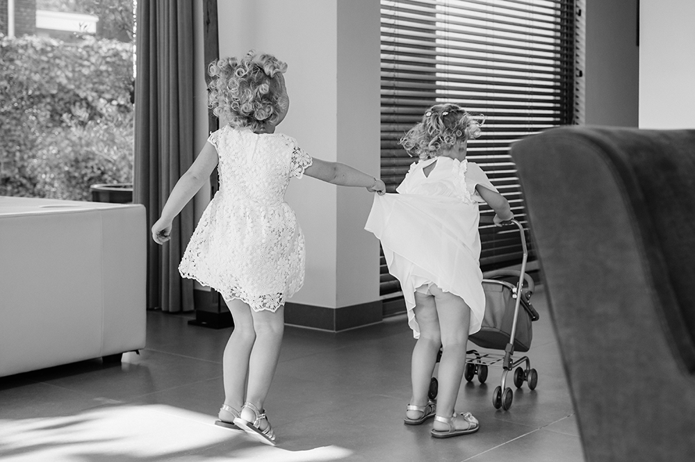 twee meisjes lopen spelend in huis waarbij het ene meisje het andere meisje haar jurk te hoog optilt-huwelijksfotografie-trouwreportage-byFotografils-IlseWagemakers-Fotograaf gevestigd in Essen