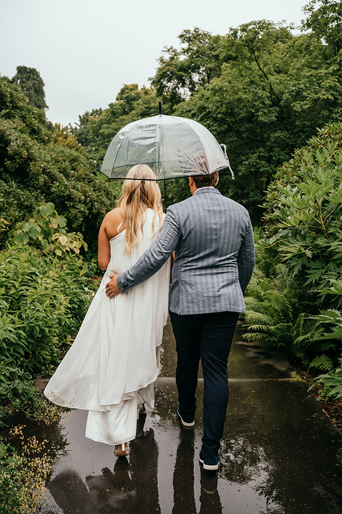 bruid en bruidegom wandelen samen onder één paraplu door het groen, foto genomen vanop de rug-huwelijksfotografie-trouwreportage-byFotografils-IlseWagemakers-Fotograaf gevestigd in Essen