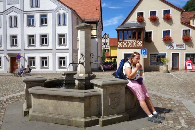 Historischer Brunnen auf einem sonnigen Platz, an dem eine Wanderin eine Pause einlegt