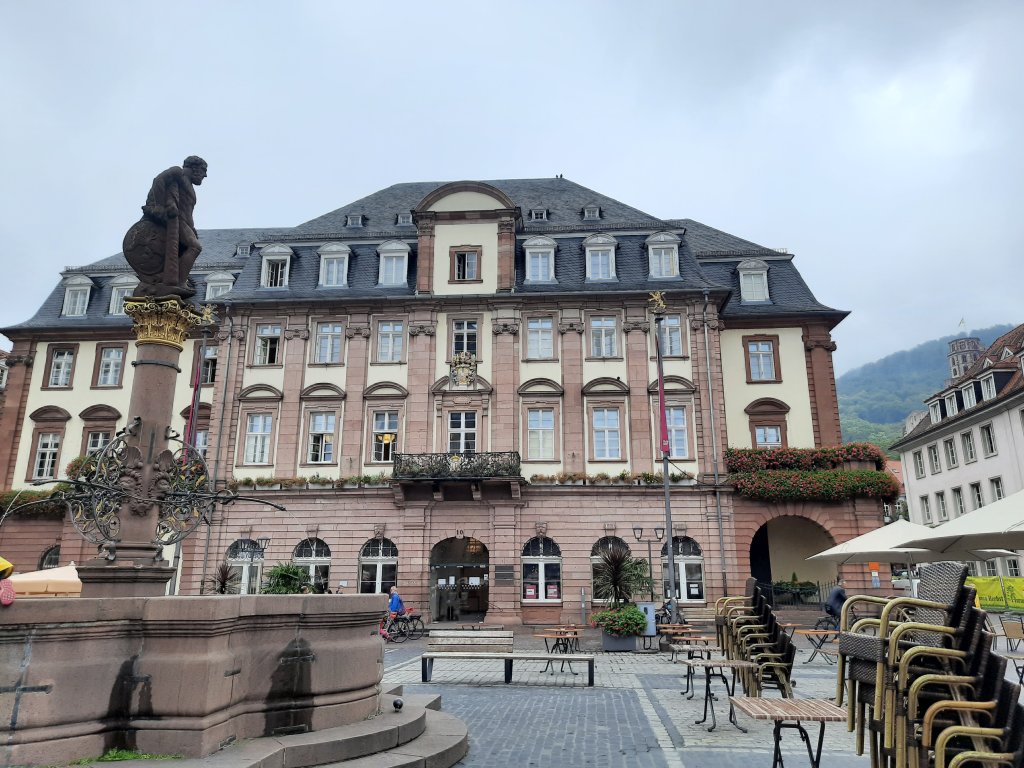 Imposantes mehrstöckiges Steingebäude am Marktplatz mit klassischer Architektur und Brunnen davor.