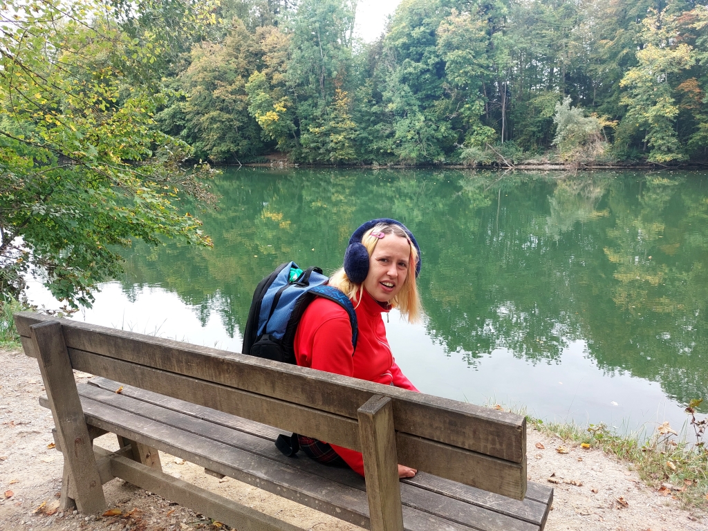 Christina mit Rucksack sitzt lächelnd auf einer Holzbank am Ufer des grünen Lechs