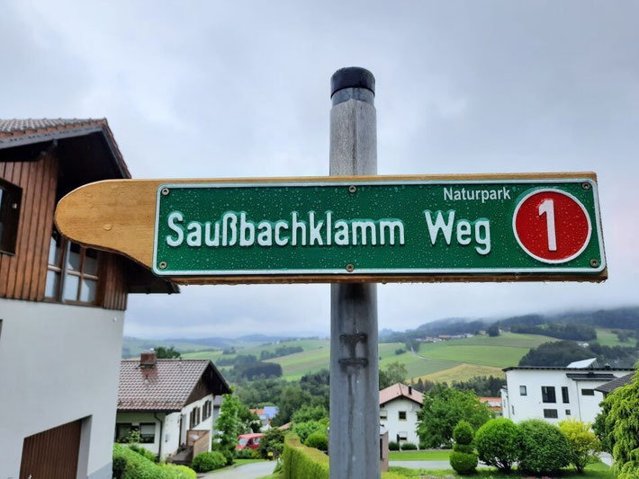 Wegweiser zum „Saußbachklamm Weg“ im Naturpark vor einer hügeligen Landschaft