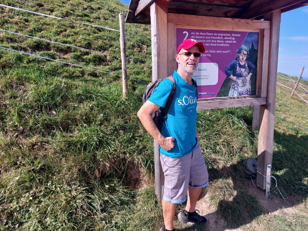 Andreas steht lächelnd vor einer Infotafel des Naturparks Nagelfluhkette auf dem Hündle