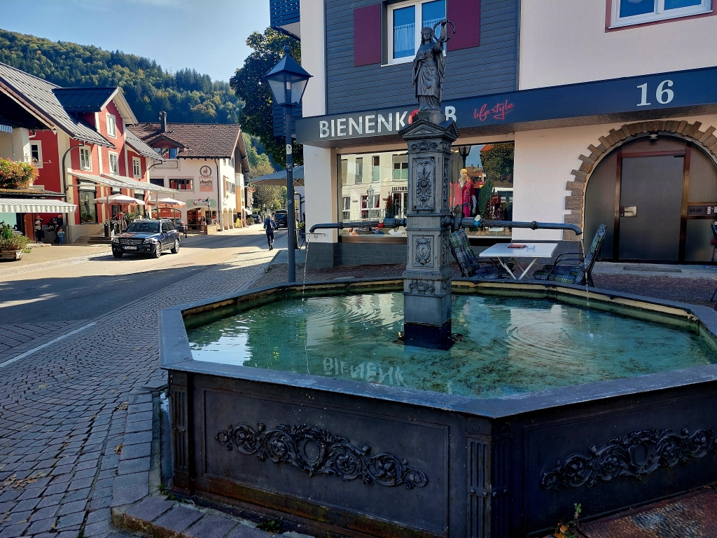 Blick auf einen Brunnen in der Ortsmitte von Oberstaufen, umgeben von gepflasterten Gehwegen
