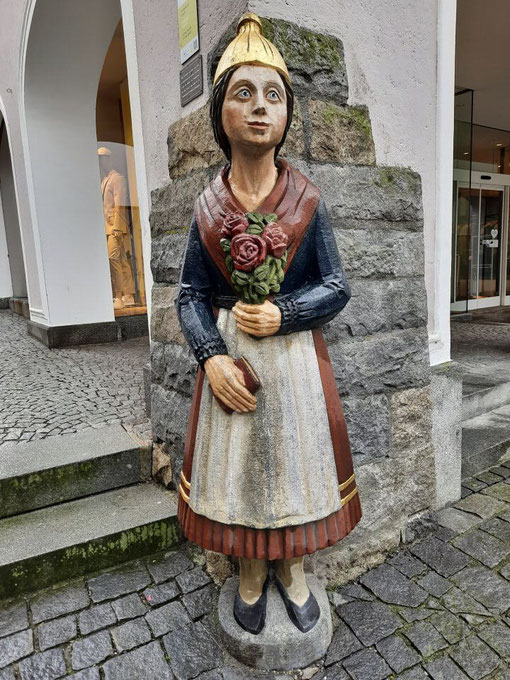 Eine dekorative Figur einer Frau in Tracht steht vor einem Gebäude im Ort.