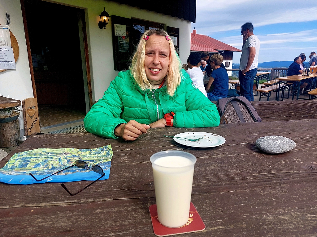 Christina sitzt bei der Einkehr an einem Holztisch vor einer Almhütte und hat ein Glas frische Milch vor sich