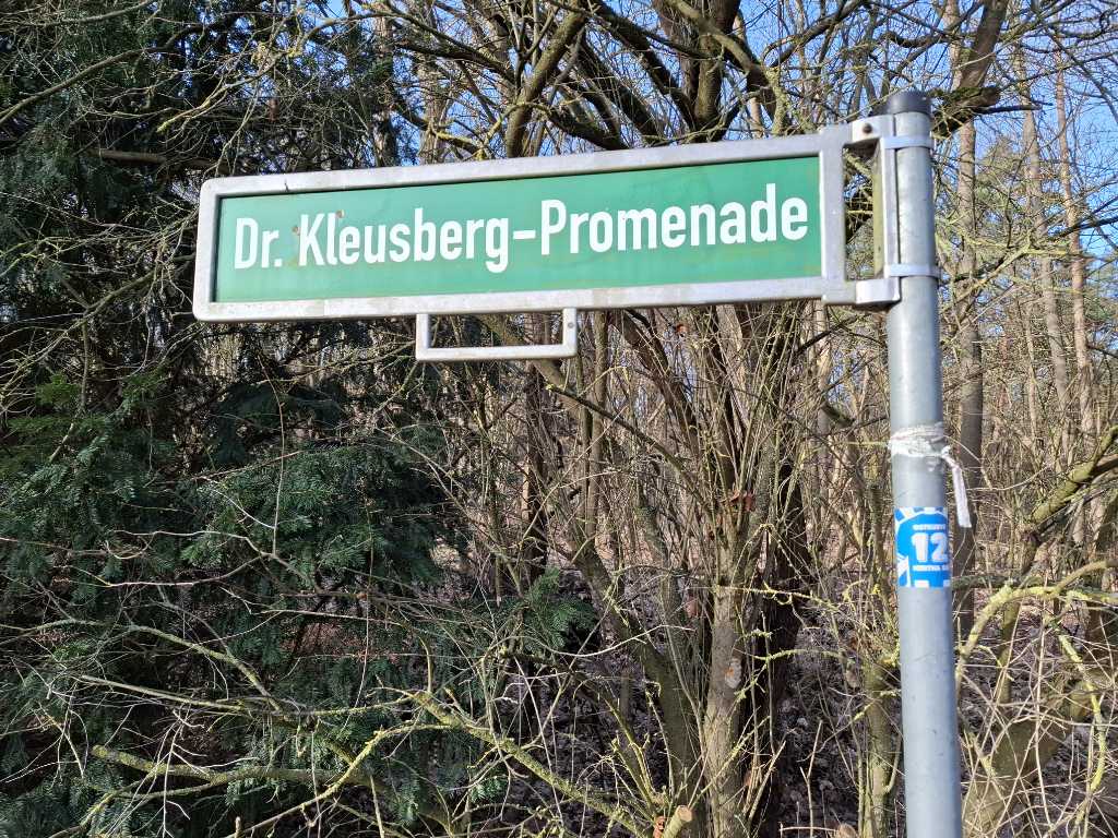 Straßenschild der Dr. Kleusberg-Promenade in Berlin-Spandau vor sonnigem Waldhintergrund