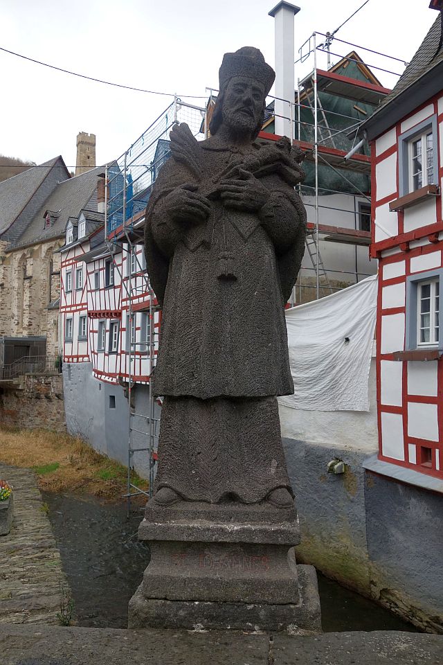 Eine steinerne Heiligenstatue (St. Johannes) auf einer Brücke vor der Kulisse des Dorfes