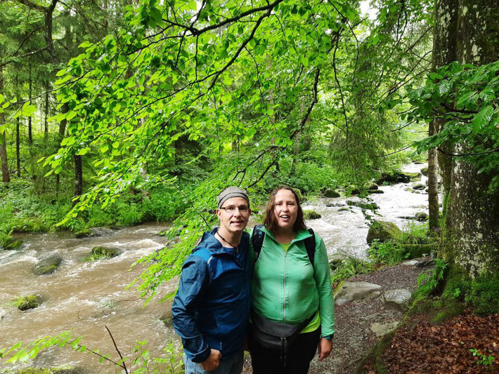 Zwei Wanderer stehen lächelnd auf einem Waldweg direkt am Wasser
