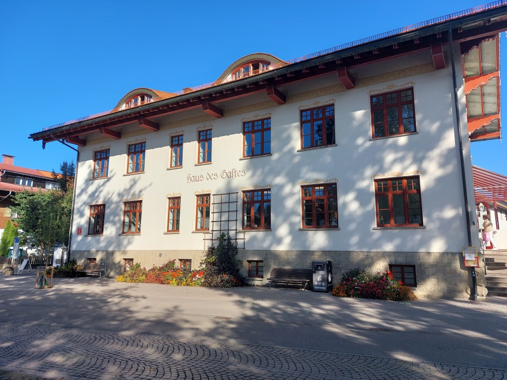 Das "Haus des Gastes" in Oberstaufen, ein großes weißes Gebäude mit markanter Holzarchitektur unter blauem Himmel