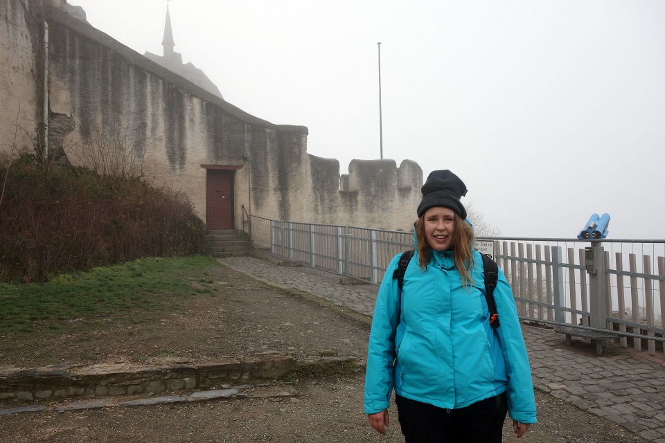 Christina lächelt in blauer Outdoor-Jacke vor den historischen Verteidigungsmauern auf dem Weg zur Burg
