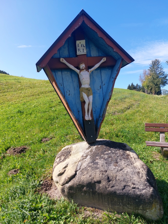 Ein blaues religiöses Denkmal (Feldkreuz) auf einem Stein am Wegrand vor Bergkulisse