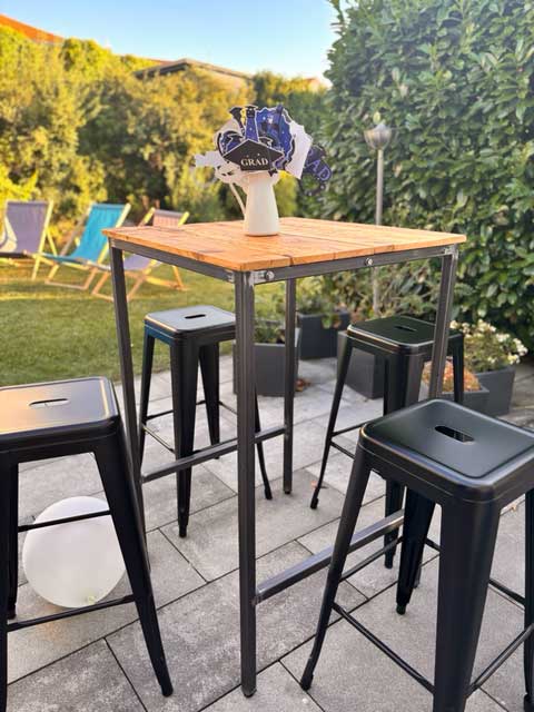 Ein Stehtisch im industriellen Design auf einer Veranstaltung mit dunkler Holzplatte und dunkelgrauen Metallfüßen. Dazu passende Hocker.