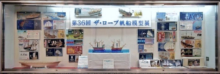 展示会場の通路側面のショーウィンドウを飾ったキットの箱絵と外輪船とスクリュウ船の綱引き動態模型。