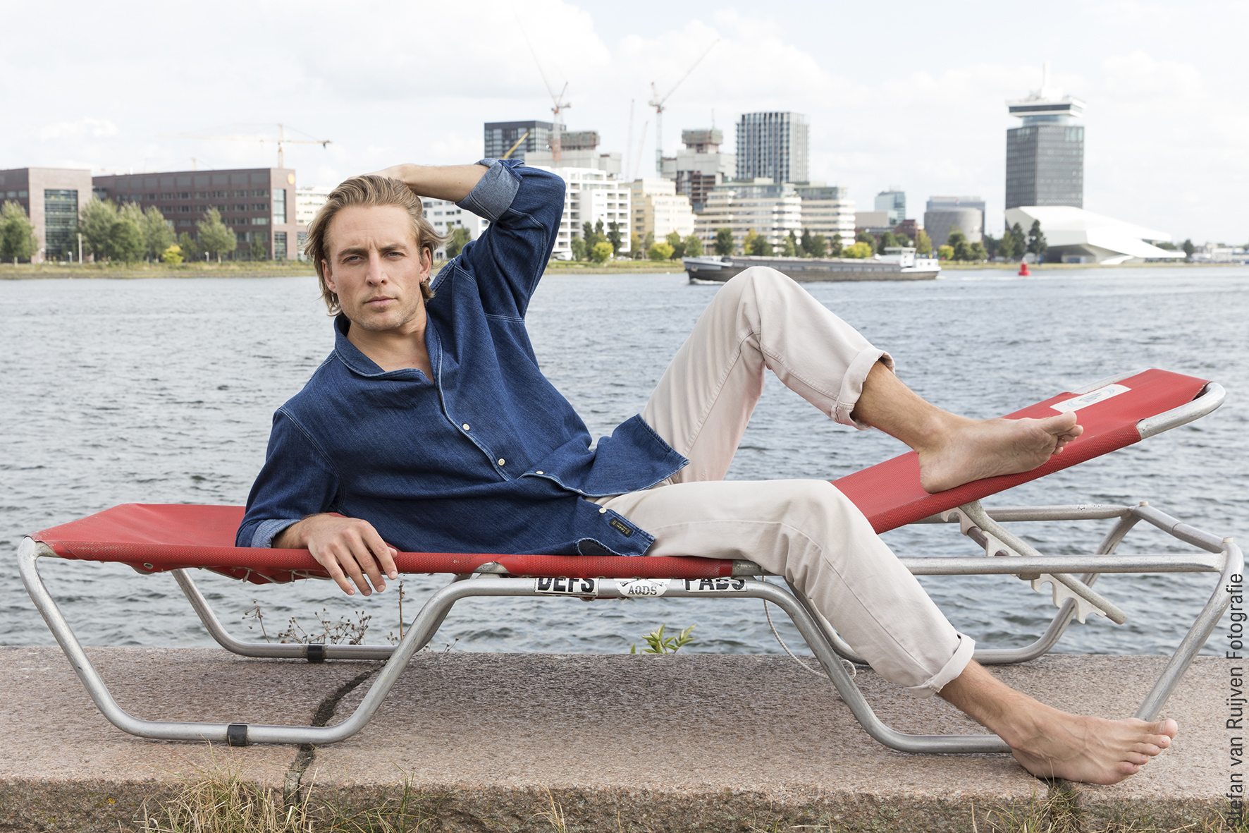 Jeroen ligt op een stretcher aan de waterkant met uitzicht gebouwen Amsterdam. Hij draagt een witte broek en denim shirt. Foto is gemaakt door Stefan van Ruijven Fotografie tijdens een portfolio update voor Jeroen.