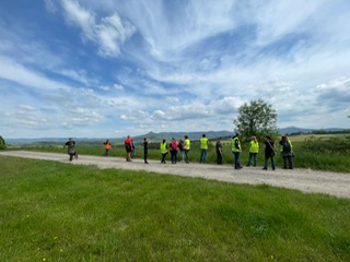 Gruppe von Personen mit Warnwesten auf einem Feldweg unter bewölktem Himmel, umgeben von Wiesen und Bäumen.