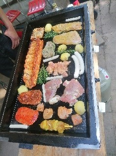 Grillplatte mit verschiedenen Fleischsorten, Würstchen, Kartoffeln und Kräutern, daneben rote Stühle und eine Person mit blauem T-Shirt.