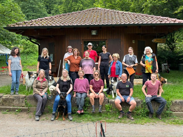 Gruppe von Menschen vor einem kleinen Holzhaus mit Ziegeldach, einige sitzen auf einer niedrigen Mauer, andere stehen, mehrere Hunde sind dabei.