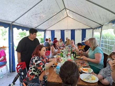 Menschen sitzen an einem langen Tisch in einem Zelt und essen gemeinsam, auf dem Tisch stehen Teller, Gläser und Flaschen.