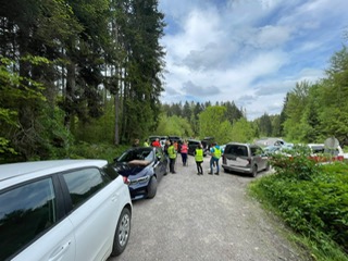 Waldweg mit mehreren parkenden Autos und einer Gruppe von Menschen in Warnwesten unter bewölktem Himmel.