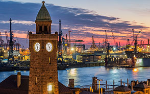 The port of Hamburg at night: It is the so-called Landungsbrücken. On the left is a large tower, in the horizontal center is water, above it many cranes.