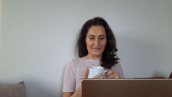 Frau lehnt mit Rücken an einer Wand mit Kaffeebecher in der Hand und Laptop vor sich