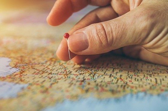 Eine Hand mit Stecknadel über einer Weltkarte bei der Urlaubsplanung.