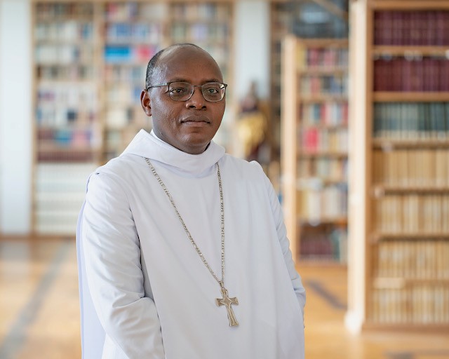 Abt Christian vor einem Raum mit Bücherregalen in einem weißen Gewand mit einem Kreuz als Halskette.