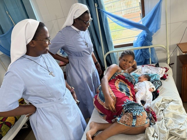 Zwei Schwestern in blauer Kleidung und eine Frau mit ihrem Baby auf einem Krankenbett. 