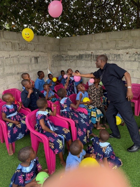 Junge Kinder sitzten im Garten auf pinken Stühlen mit bunten Luftballons, Betreuer steht vor den Kindern und lüpft mit der Hand einen Ballon in die Luft.
