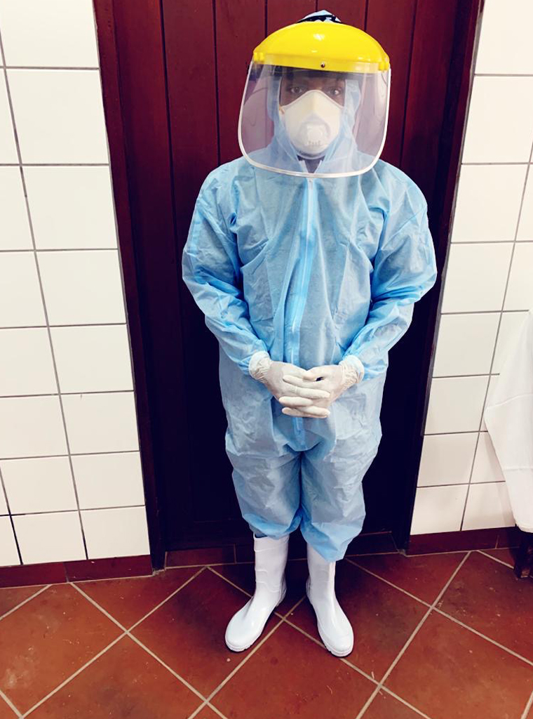 Pflegekraft in vollständiger Corona-Schutzausrüstung mit Overall, Maske, Visier und Überschuhen steht vor einer Tür.