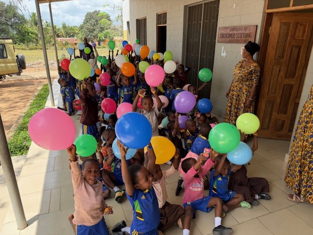 Kleine Schulkinder sitzen und stehen vor Klassenzimmer mit bunten Luftballons und freuen sich.
