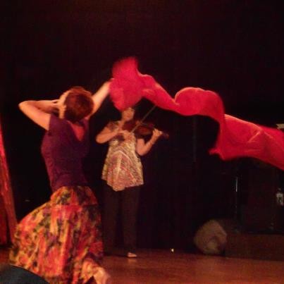 A dancer is kneeling with red silk flowing above her. A violinist stands in the background, playing over the woman as she worships.