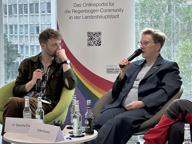 Zwei Personen sitzen auf grünen Sesseln bei einer Podiumsdiskussion: Eine hält ein Mikrofon. Im Hintergrund hängt ein Banner mit der Aufschrift Das Onlineportal für die Regenbogen-Community in der Landeshauptstadt.