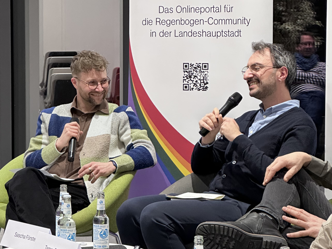 Zwei lächelnde Männer führen eine moderierte Diskussion auf der Bühne vor einem Banner mit der Aufschrift 'Das Onlineportal für die Regenbogen-Community in der Landeshauptstadt'.