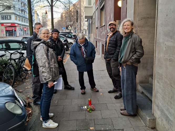 Gruppe von sieben Personen steht auf einem Bürgersteig in einer städtischen Umgebung; im Vordergrund sind Blumen und eine Kerze als Gedenkzeichen auf dem Boden zu sehen.