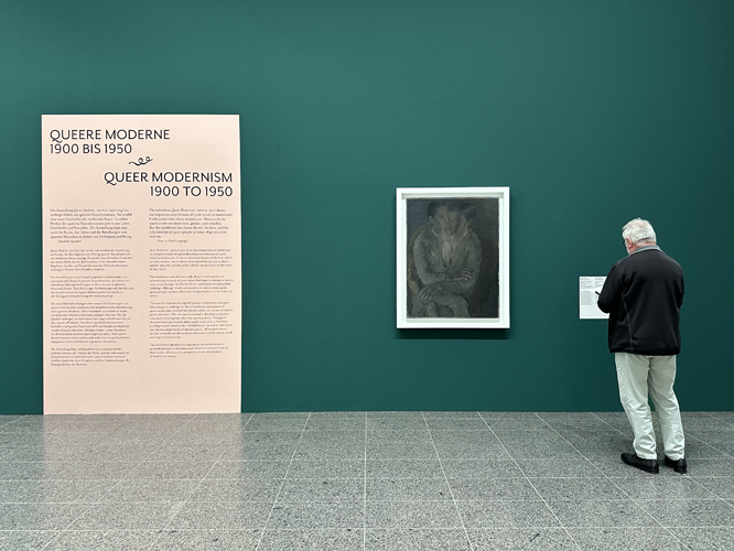 Besucher betrachtet in einer Ausstellung ein Gemälde an einer dunklen Wand, daneben eine Infotafel zur Ausstellung 'Queere Moderne 1900 bis 1950'.