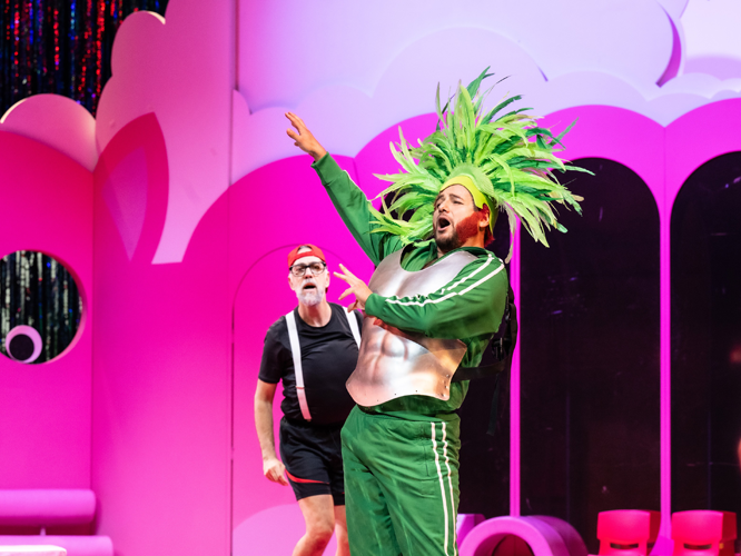 Zwei Schauspieler auf einer Bühne: Einer trägt ein grünes Kostüm mit Federschmuck und posiert singend, der andere steht im Hintergrund in einem schwarzen Outfit mit roten Accessoires.