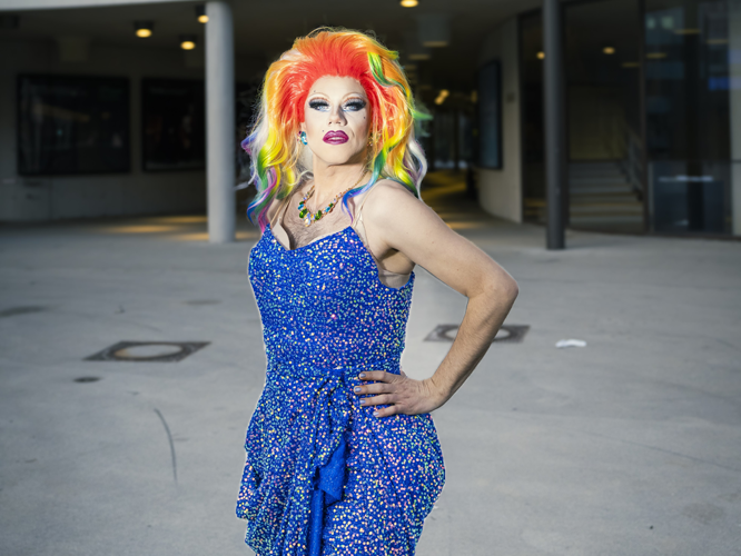 Eine Person mit buntem, rainbow-farbenem Haar in einem blauen, gemusterten Kleid posiert selbstbewusst mit einer Hand an der Hüfte vor einem modernen Gebäude.