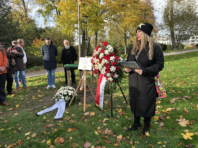 Eine Person hält eine Rede bei einer Gedenkveranstaltung im Freien. Im Vordergrund stehen zwei Kränze mit Blumen, einer davon mit roten und weißen Blüten.