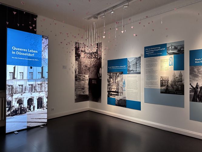 Ausstellungsraum mit der Ausstellung ‚Queeres Leben in Düsseldorf‘, die historische Fotos, Informationstafeln und künstlerische Installationen zeigt.