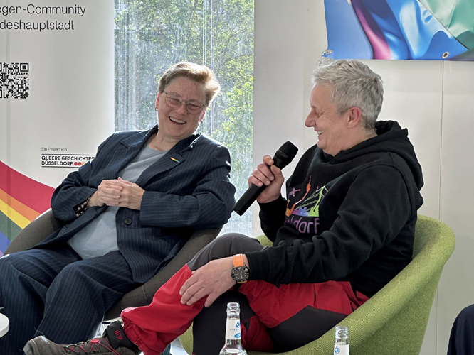 Zwei Personen sitzen auf grünen Sesseln bei einer Podiumsdiskussion: Eine hält ein Mikrofon, die beiden unterhalten sich lachend.